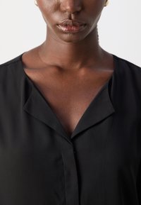 Blusa nera con scollo a V profondo e una leggera lucentezza. Presenta una texture liscia e un dettaglio con un solo bottone all'apertura del collo.