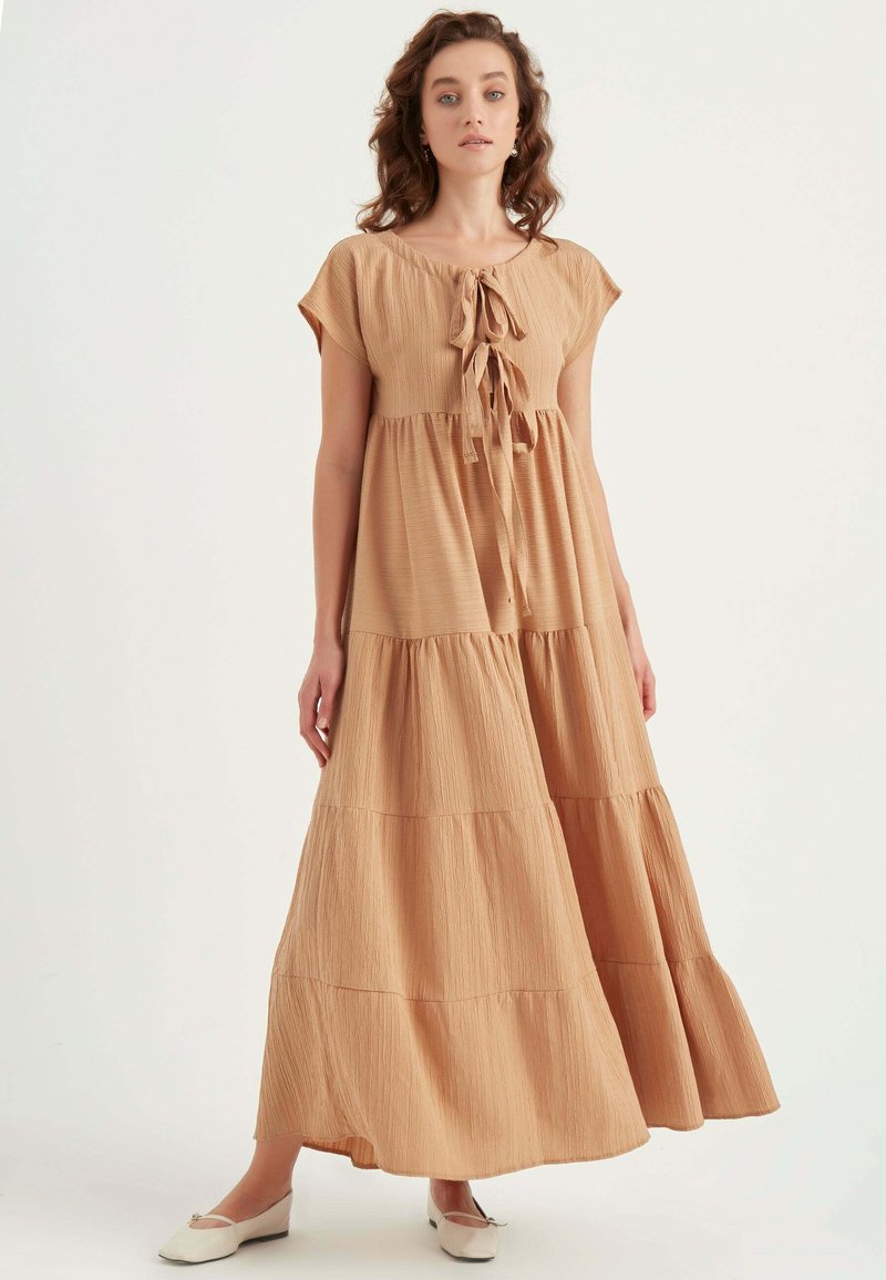 Langes, gestuftes Kleid aus weichem braunem Stoff mit strukturierter Oberfläche. Hat kurze Ärmel und einen Bindeverschluss am Ausschnitt.
