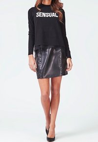 Suéter negro con el texto "SENSUAL", combinado con una mini falda de cuero negro. La falda tiene una textura suave y brillante. Unos tacones altos negros completan el look.