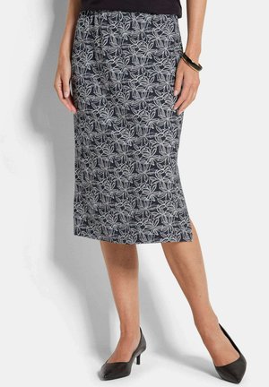 Femme portant une jupe noire mi-longue à motifs floraux blancs, des chaussures à talons noires et un bracelet noir au poignet droit.