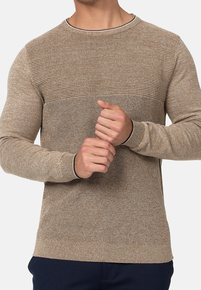 Homme portant un pull en tricot beige avec un motif côtelé sur la poitrine et des manches en tricot uni, joignant ses mains devant lui.