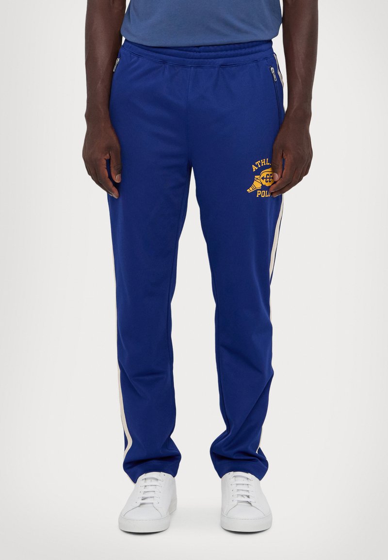 Pantaloni sportivi blu con logo dorato, vita elastica, tasche con zip e strisce laterali bianche, realizzati in tessuto morbido e leggero.
