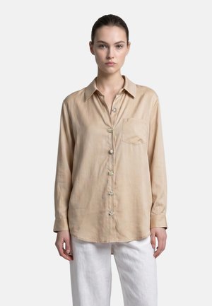 Young woman wearing a beige long-sleeve button-up shirt with decorative buttons and white pants standing against a plain background.