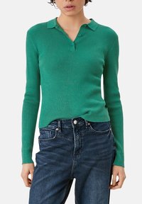 Grünes, geripptes Langarm-Polohemd mit Kragen und drei Knöpfen, kombiniert mit dunkelblauen Jeans.