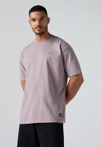 T-shirt en coton violet clair à manches courtes, à col rond, avec un petit logo sur la poitrine ; associé à un pantalon noir.
