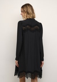 Schwarzes Kleid mit langen Ärmeln, das mit Spitzenakzenten entlang des Rückens und des Saums versehen ist. Weicher Stoff mit lockerer Passform, der oberhalb des Knies endet.