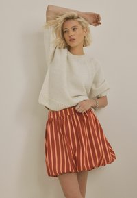 Pulover ușor, tricotat, cu model texturat, în nuanță de crem, asortat cu o fustă plisată cu talie înaltă, prezentând dungi verticale roșii și portocalii.