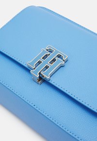 Světle modrá strukturovaná kožená kabelka s příklopem, na kterém je stříbrný a modrý dvojitý T logo klip.