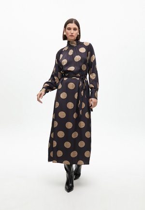 Vestido de manga larga azul marino con grandes lunares beige, cuello alto, cintura ceñida y tejido fluido. Combinado con botas negras hasta la rodilla.