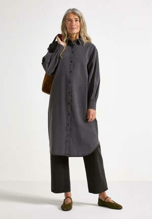 Abito camicia in denim grigio con chiusura a bottoni, maniche lunghe e aperture laterali. Abbinato a pantaloni larghi neri e scarpe verde oliva.