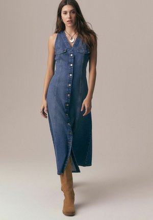 Kobieta ubrana w bezrękawnikową, zapinaną na guziki sukienkę z denimu z kieszeniami na piersi i beżowe kozaki za kolano, stojąca na tle gładkiego tła.