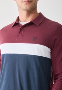 Langermet polo skjorte med en maroon topp, hvit midtstripe og marineblå bunnseksjon. Laget av myk stoff med et brodert logo.