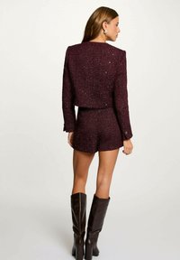 Completo bordeaux in due pezzi composto da una giacca corta a maniche corte, texturizzata e scintillante, e pantaloni abbinati, abbinato a stivali marroni alti fino al ginocchio.
