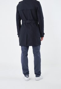 Marinblå trenchcoat med bältad midja och axelremmar, kombinerad med blå byxor och vita sneakers. Enkel design, mjuk tyg.