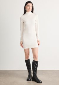 Rochie tricotată de culoare crem, cu guler înalt și mâneci lungi, completată de cizme negre până la genunchi cu fermoar lateral și finisaj texturat.