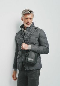 Szara ocieplana kurtka z wysokim kołnierzem, guzikami i dwiema kieszeniami, zestawiona z czarną, teksturowaną torbą na ramię. Ciemne dżinsy i szara bluza pod spodem.