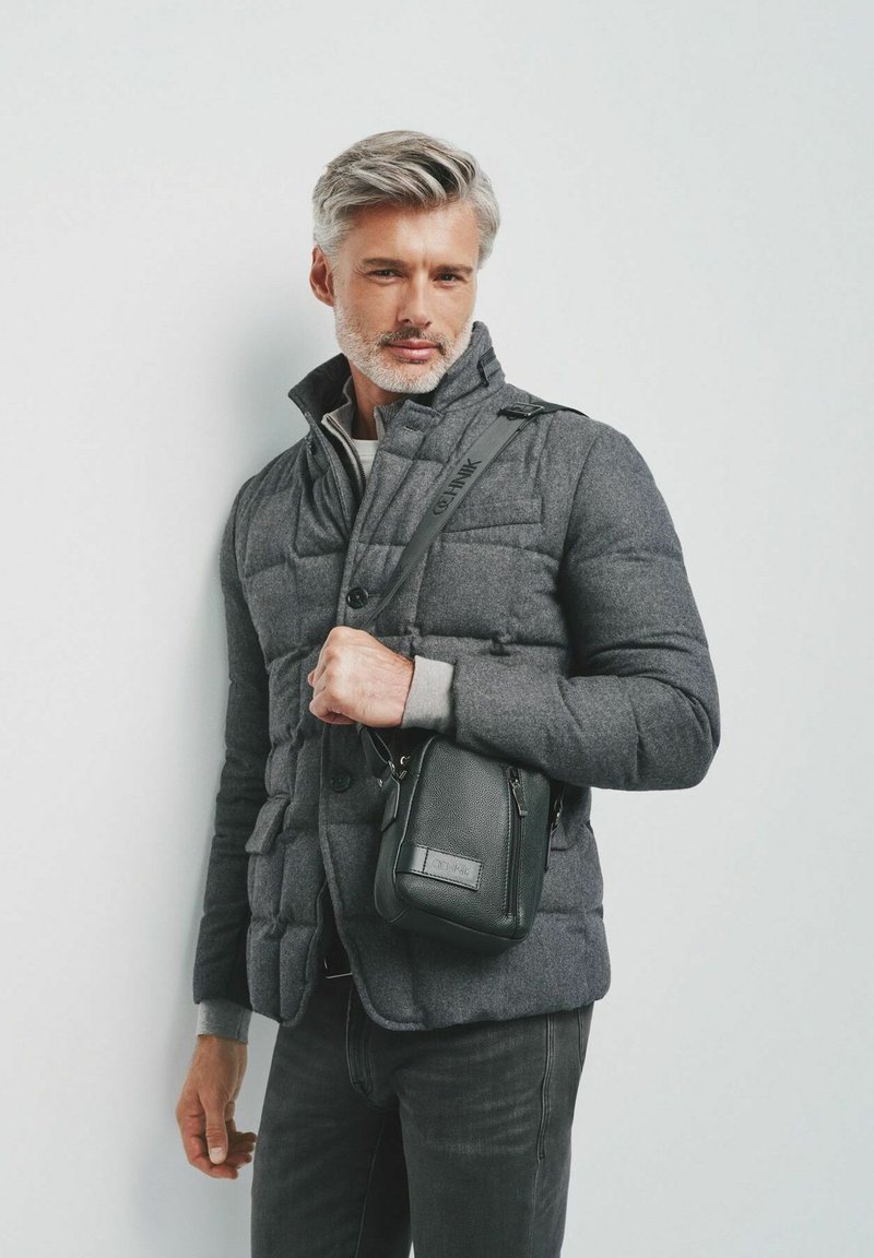 Szara ocieplana kurtka z wysokim kołnierzem, guzikami i dwiema kieszeniami, zestawiona z czarną, teksturowaną torbą na ramię. Ciemne dżinsy i szara bluza pod spodem.