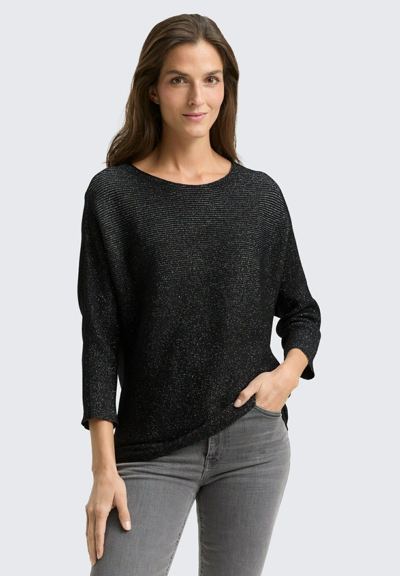 Pull en maille métallique noir à coupe décontractée, col bateau et manches trois-quarts. Surface texturée avec un léger éclat, porté sur un jean gris.