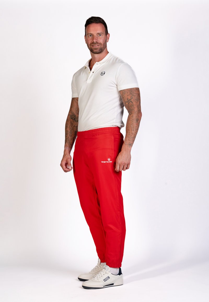Rode, elastische joggingsbroek met een logo op de linker dij, gecombineerd met een getailleerd wit poloshirt en witte sneakers. Zachte textuur.