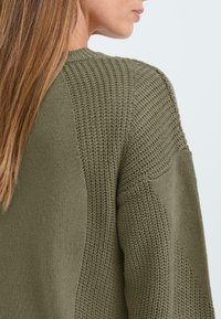 Olivgrön stickad tröja med texturerade paneler, ribbade detaljer och avslappnad passform, som visar en blandning av stickmönster och slät tyg.