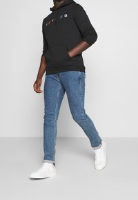 Sudadera negra con el texto "NOTHING" multicolor, combinada con vaqueros ajustados azules y zapatillas blancas, mostrando un estilo casual y moderno.
