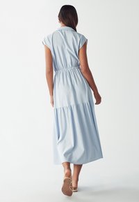 Abito maxi azzurro chiaro con maniche a cappuccio, vita aderente e gonna a strati. Tessuto morbido, la sartoria valorizza la silhouette.