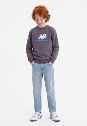 Smilende rødhåret dreng iført mørkegrå New Balance sweatshirt, lyseblå jeans og hvide sneakers med blå accenter, hænderne i lommerne.