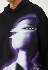 Svart hettegenser med et frontgrafisk design i nyanser av lilla og hvitt. Teksten "HYPERSPACE" er trykt i en fet, moderne skrifttype.