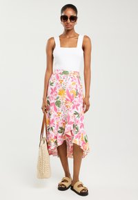 Haut blanc à col carré, jupe fleurie rose avec ourlet volanté, portée avec des sandales beiges et tenant un sac en osier beige. Lunettes de soleil rondes foncées.