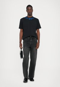 Svart t-shirt med blå text längs halsringningen, kombinerad med svarta jeans och hållande en liten svart väska. Platta skor fulländar looken.