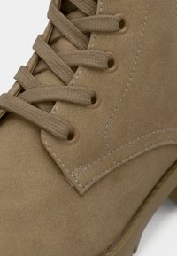 Gros plan d’une botte beige en daim à lacets montrant des lacets en tissu texturé, des œillets métalliques et des coutures détaillées sur la partie supérieure de la chaussure.