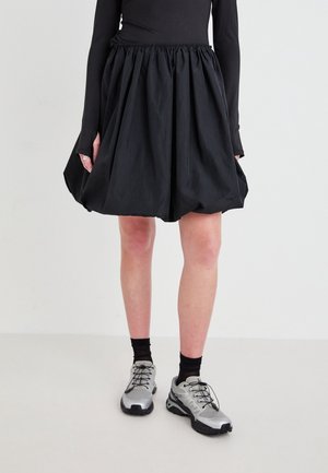 Jupe noire mi-longue avec taille élastique, silhouette bouffante, confectionnée dans un tissu léger. Associée à des chaussures de sport grises et des chaussettes noires.