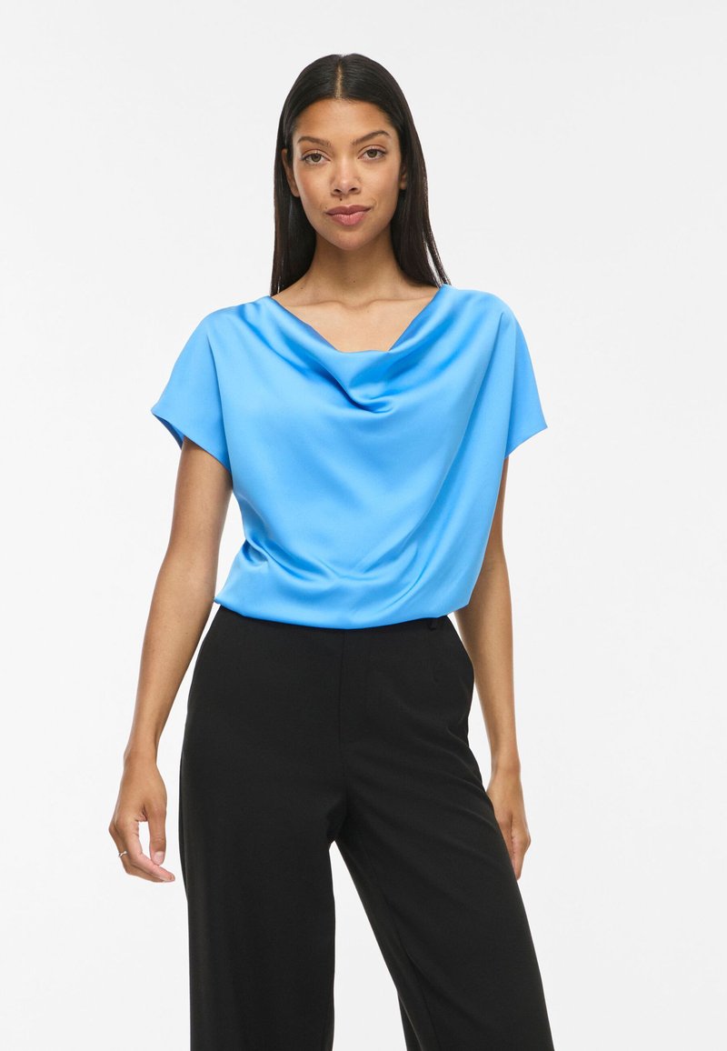 Femme aux longs cheveux foncés portant un chemisier bleu vif drapé à manches courtes et un pantalon noir taille haute, debout sur un fond blanc.
