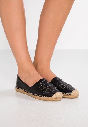 Füße tragen schwarze Slip-On-Espadrilles mit beigen, gewebten Zehenkappen und genähten Sohlen, mit großem geprägt Logo oben.