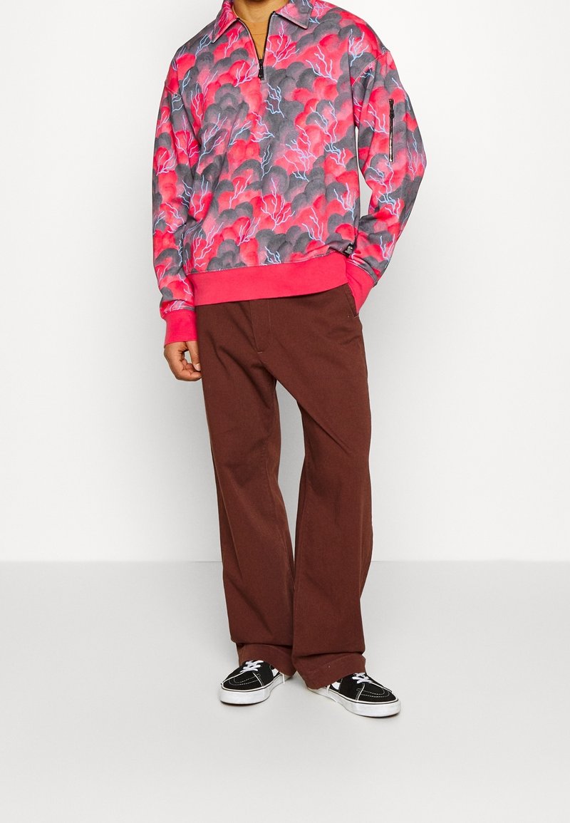 Personne portant un sweat à capuche zippé rose et gris à motifs, un pantalon large marron et des baskets noires et blanches, debout sur un fond blanc.