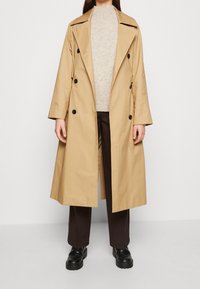 Gabardina beige de doble botonadura con grandes botones negros, solapas amplias y mangas largas, superpuesta a un suéter claro y pantalones oscuros.