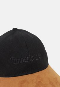 Černá textilní čepice s hnědým semišovým šiltovým okrajem, na přední straně s embosovaným logem "Timberland". Viditelné švy podél koruny.