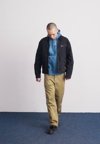 Black jacket over a blue hoodie, beige pants, and black shoes. The outfit features a front zipper and a casual, layered design.