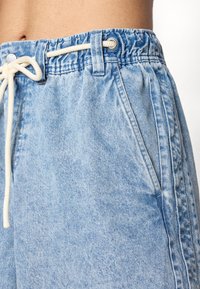 Lyseblå denimshorts med elastisk talje og hvid snor, båret af en person med synlig hud på maven.
