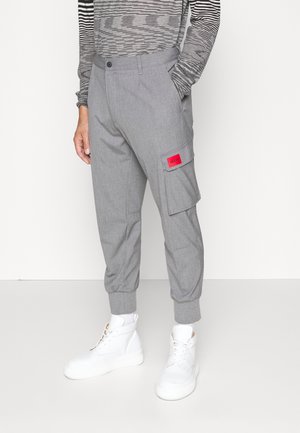Persoon die grijze cargobroek draagt met een rode patch op de zijzak, een gestreept shirt met lange mouwen, witte sokken en witte hoge sneakers.
