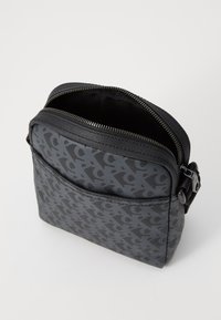 Geantă de umăr din material textil negru cu un model texturat. Dispune de un sistem de închidere cu fermoar și o buzunare exterioară pe față. Formă compactă, pătrată.