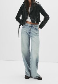 Chaqueta de motorista de cuero negro, top corto blanco y vaqueros de pierna ancha color azul claro. Detalles notables incluyen cintura alta y textura de denim desgastado. Zapatos negros.