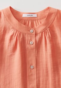 Camicia color pesca con scollatura rotonda, dettaglio plissettato e tre bottoni bianchi sul davanti. Tessuto morbido e strutturato.