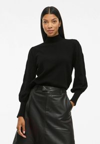 Femme aux longs cheveux noirs portant un pull à col montant noir avec des manches bouffantes et une jupe en cuir noir, se tenant avec une main dans la poche.