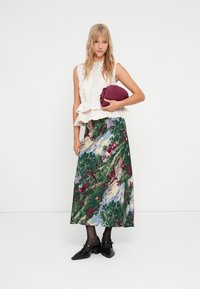 Hvit peplum-top, lang flerfarget blomsterkjole med landskapsprint, holder en bordeaux clutch, svarte strømper og svarte sko.