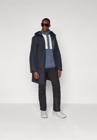 Veste imperméable marine avec une capuche, design en blocs de couleur gris clair, associée à un pull bleu et un pantalon noir. Baskets blanches et lunettes de soleil.