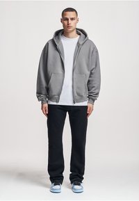 Grauer Kapuzenpullover mit Reißverschluss, großer Kapuze und Fronttaschen, getragen über einem weißen T-Shirt, kombiniert mit schwarzen Hosen und Sneakern mit blauen Akzenten.