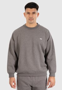 Sweat-shirt gris en tissu doux, avec un col rond et des poignets côtelés. Un petit logo blanc est visible sur le devant.