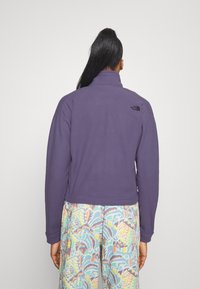 Fioletowa polarowa kurtka z wysokim kołnierzem i małym czarnym logo na górnej części pleców. Model nosi wzorzyste, kolorowe spodnie.