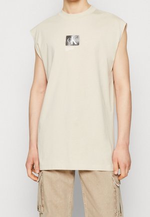 T-shirt sans manches en coton beige avec un logo "CK" carré en noir et argent imprimé sur le devant. Coupe droite.